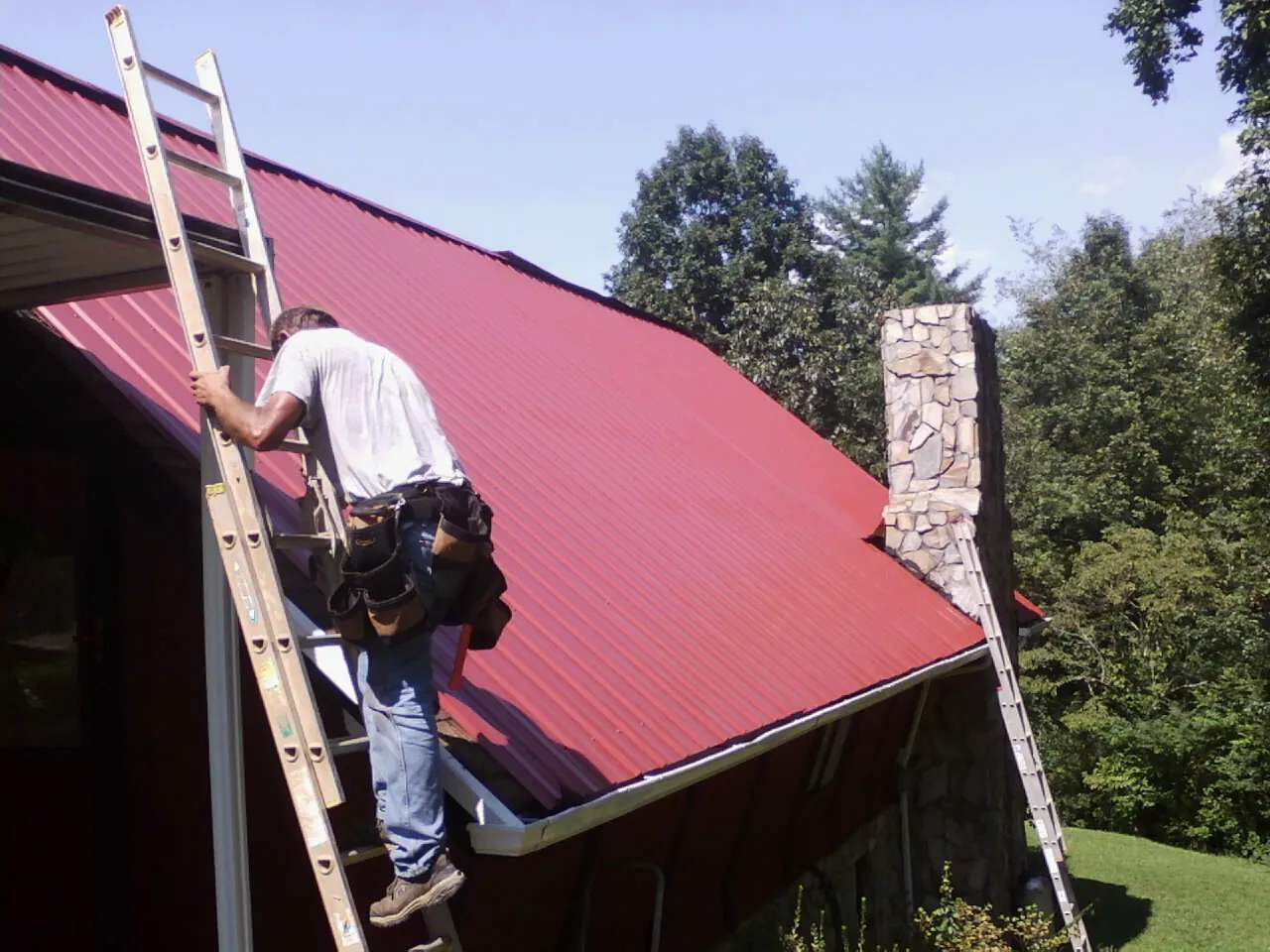 Roofer on a ladder beside a red metal roof and stone chimney — Soffit & Fascia Repair by Roofing Brothers in Royse City